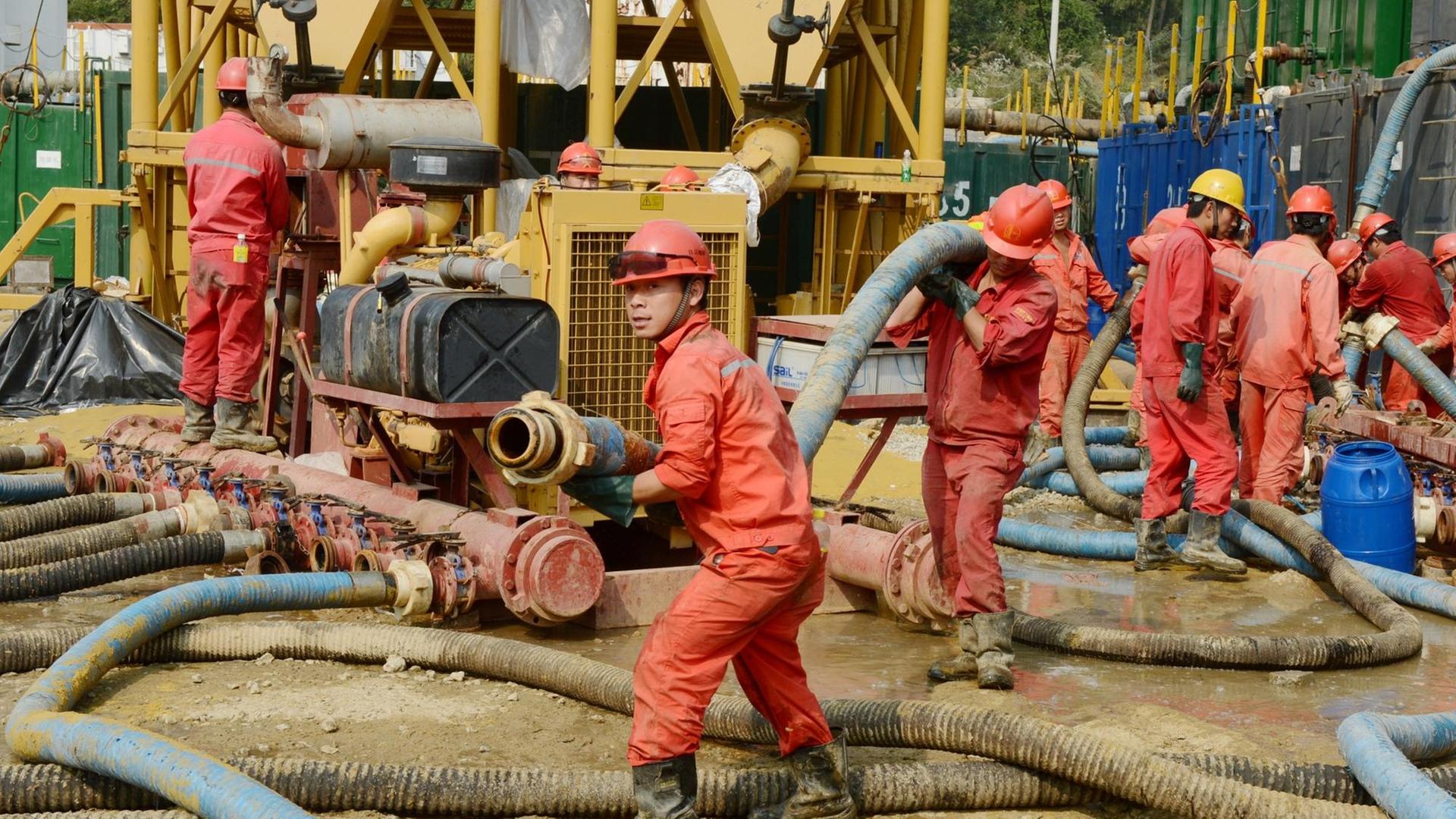 Chinesische Arbeiter in roten Overalls tragen armdicke Rohre für eine Schiefergasanlage durch den Matsch in Zentralchina.