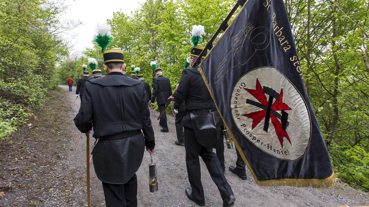 Bergleute in traditioneller Uniform gehen einen Weg entlang, mit dem Rücken zum Betrachter. Einer der Männer trägt eine schwarze Fahne, auf der Grubenwehr Prosper-Haniel steht.