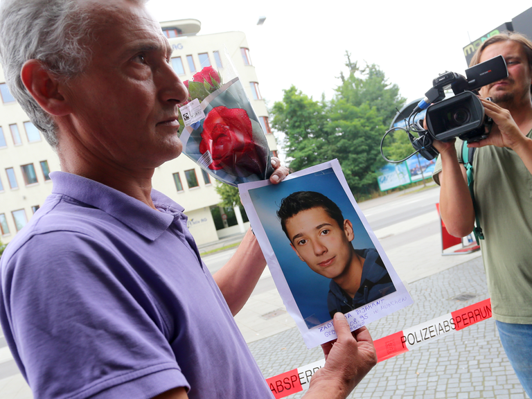 Naim Zabergja steht mit einem Foto seines getöteten Sohnes Dijamant am 23.07.2016 vor Medienvertretern am Olympia-Einkaufszentrum in München (Bayern), das die Polizei nach einer Schießerei mit Toten und Verletzten am Vortag abgesperrt hat.