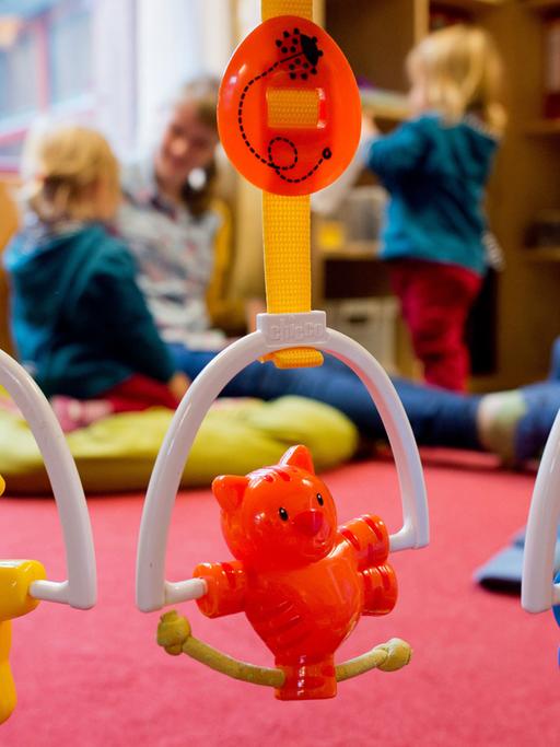 Kinderspielzeug hängt an einem Rahmen. Im Hintergrund spielt eine Erzieherin mit zwei kleinen Kindern.