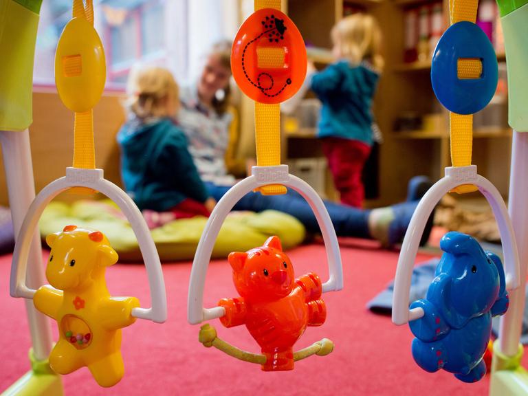 Kinderspielzeug hängt an einem Rahmen. Im Hintergrund spielt eine Erzieherin mit zwei kleinen Kindern.
