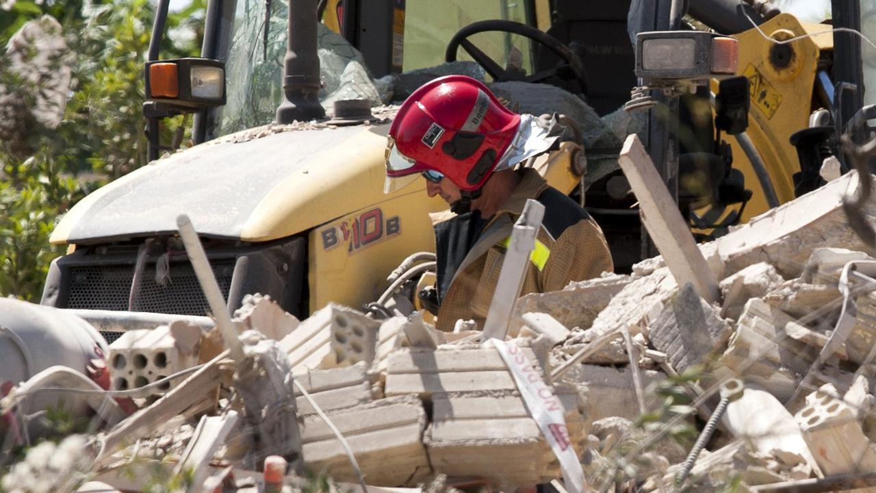 Der Ermittler mit rotem Helm und Sonnenbrille steht vor einem gelben Traktor und hinter einem Trümmerhaufen.