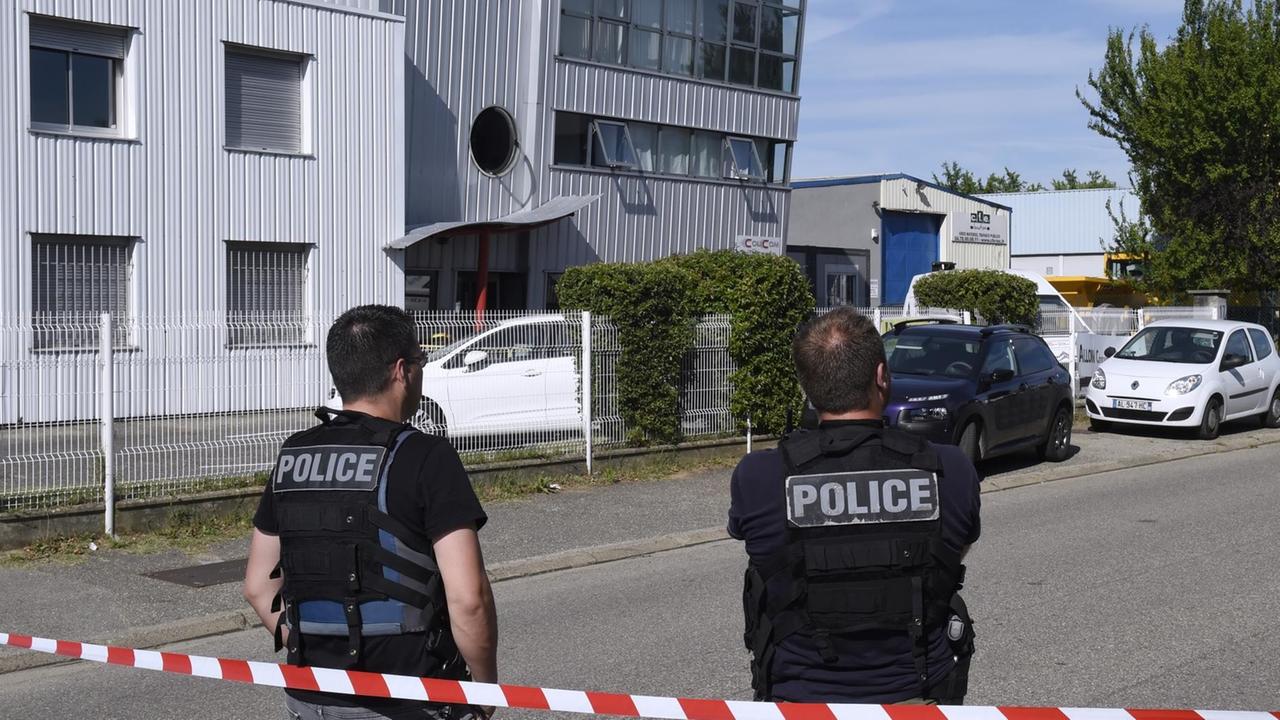 Zwei Polizisten stehen hinter einem weiß-roten Absperrband auf der Straße vor der Lieferfirma. Auf ihren Rücken steht "Police". Zwei Polizisten stehen hinter einem weiß-roten Absperrband auf der Straße vor der Lieferfirma. Auf ihren Rücken steht "Police".