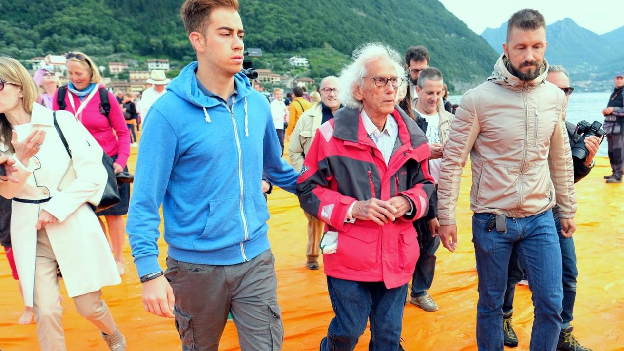 Künstler Christo (M) wird am 18.06.2016 vor Paratico auf dem Lago d'Iseo in Italien von Begleitern durch die Besuchermassen über die schwimmendenen Stege mit orangefarbenen Stoff seines Projekts "The Floating Piers" geführt.