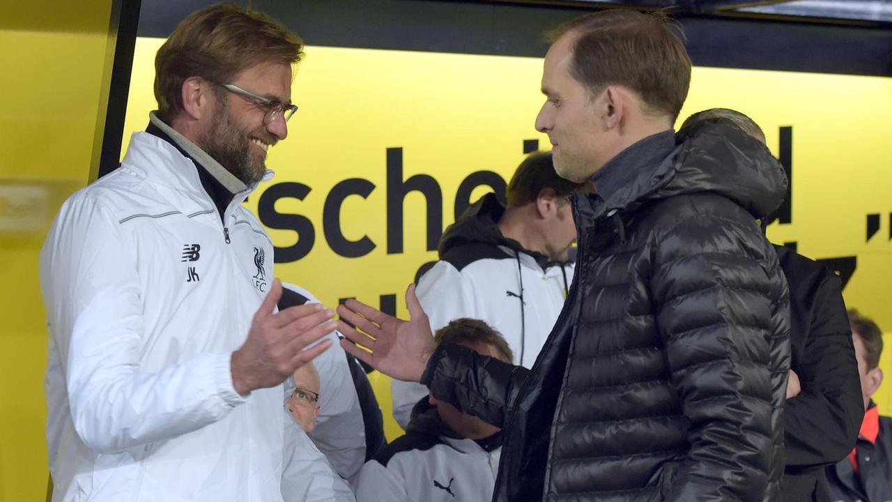 Klopp in weißer Sportjacke und Tuchel in einer schwarzen Winterjacke geben sich vor einer Ersatzbank lächelnd die Hand. Klopp in weißer Sportjacke und Tuchel in einer schwarzen Winterjacke geben sich vor einer Ersatzbank lächelnd die Hand.