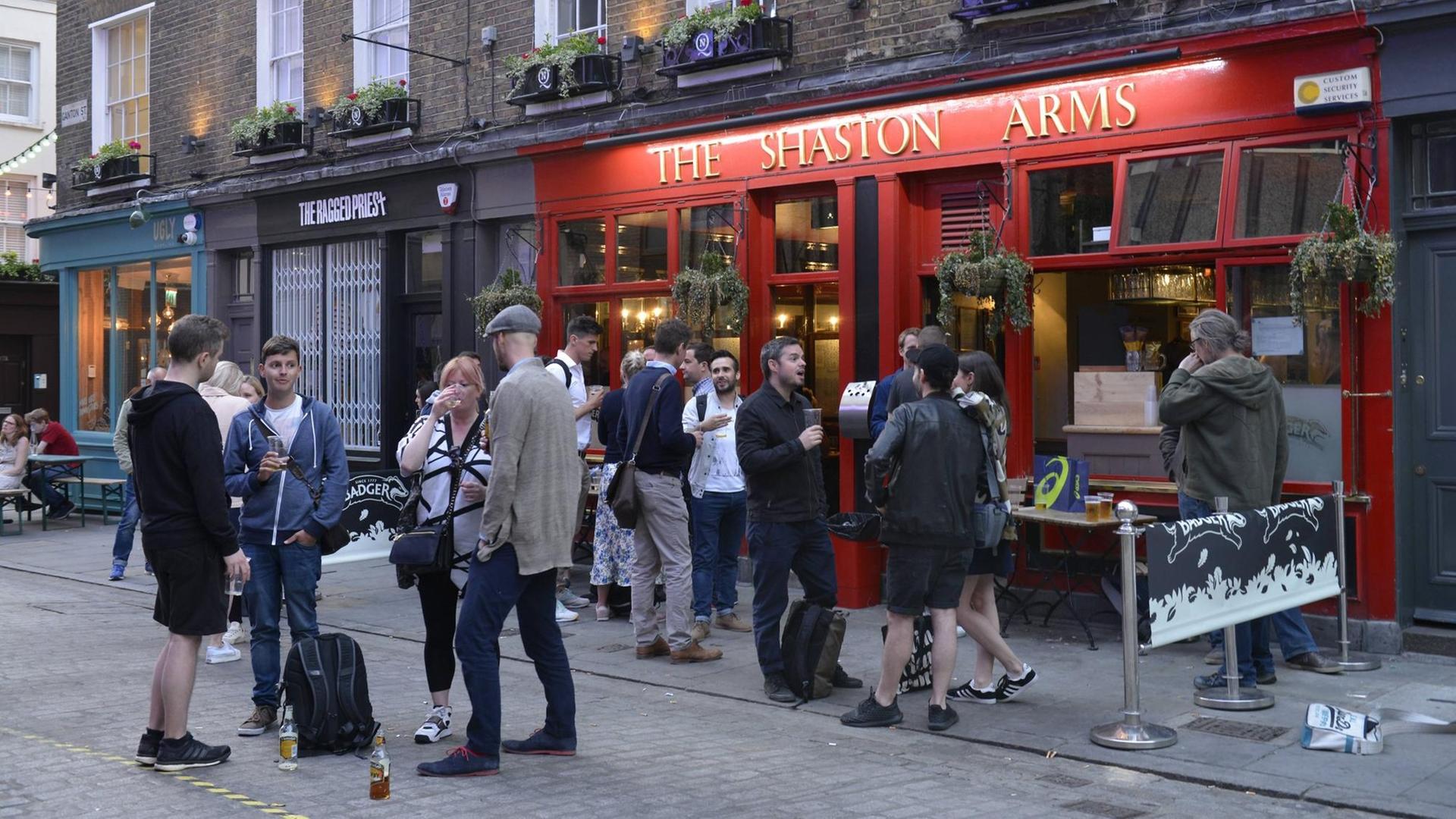 Sie sehen einen Pub in London von außen, viele Menschen stehen davor. Sie sehen einen Pub in London von außen, viele Menschen stehen davor.