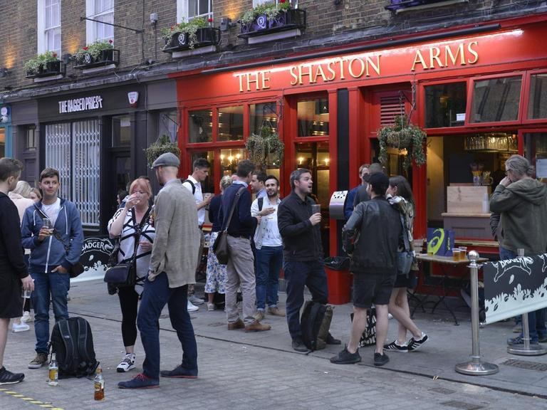 Sie sehen einen Pub in London von außen, viele Menschen stehen davor.
