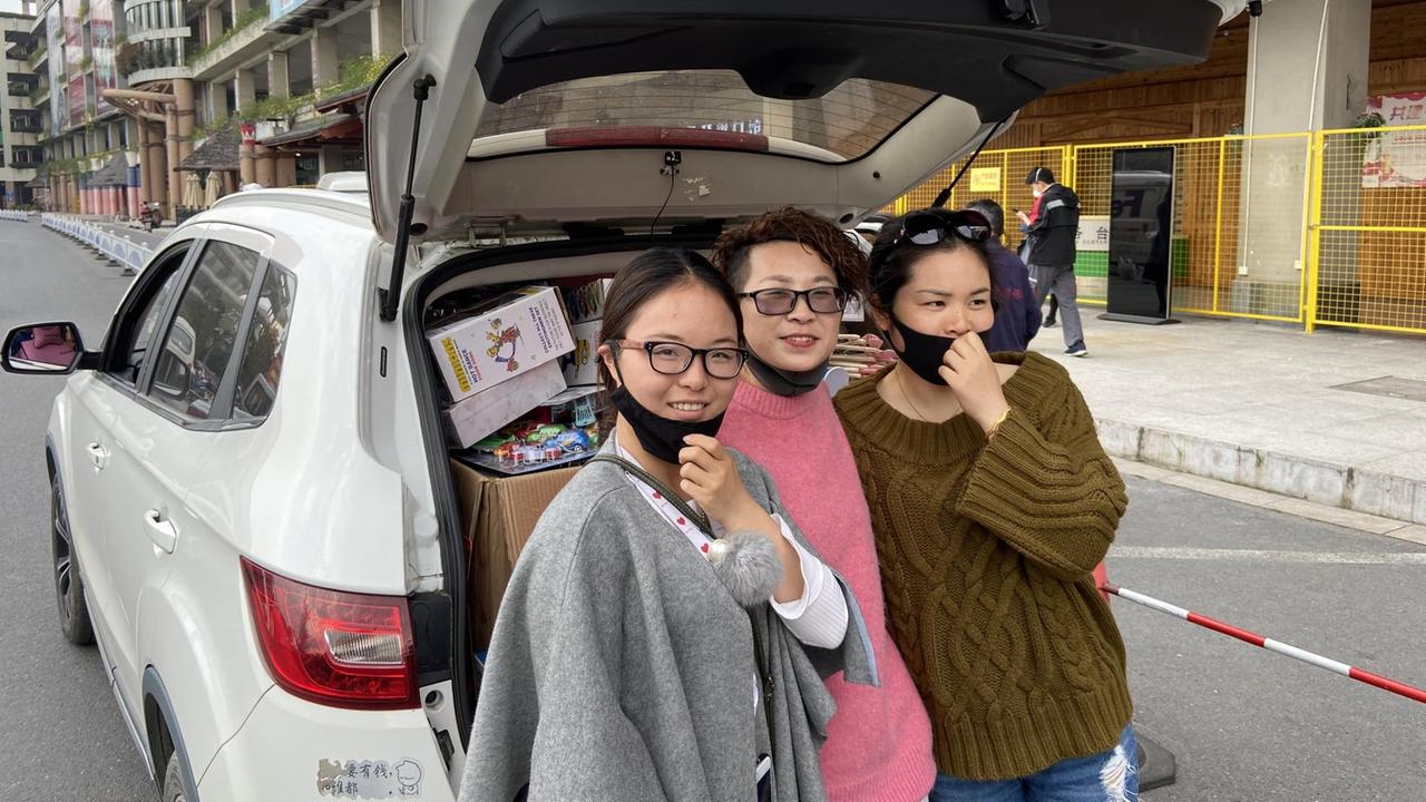 Drei junge Frauen stehen nebeneinander vor dem geöffneten Kofferraum eines Autos.