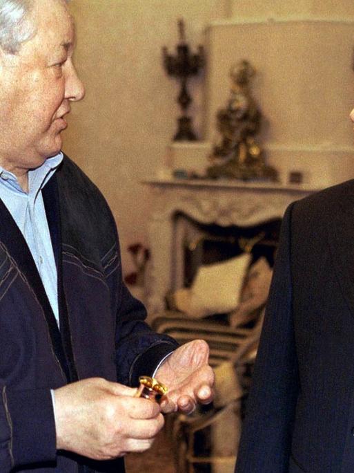 Präsident Putin steht rechts in einem noblen Innenraum udn schaut den älteren Boris Jelzin von der Seite erwartungsfroh an.
