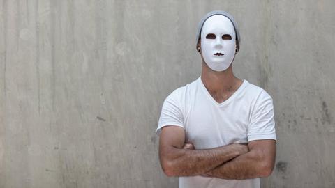 Ein Mann mit weißer Maske und verschränkten Armen steht vor einer Betonwand