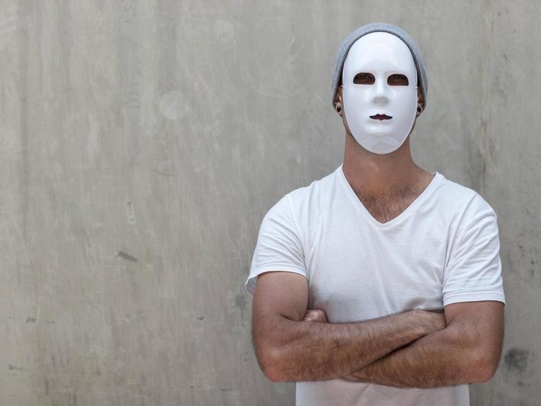 Ein Mann mit weißer Maske und verschränkten Armen steht vor einer Betonwand