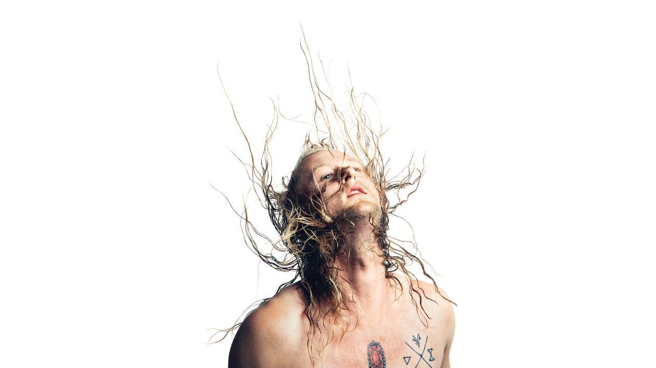 Auf dem Bild ist der Musiker Colin Self zu sehen. Er wirbelt seine nassen Haare in die Luft. Sein Oberkörper ist nackt. Auf seiner Brust hat er zwei Tätowierungen. 