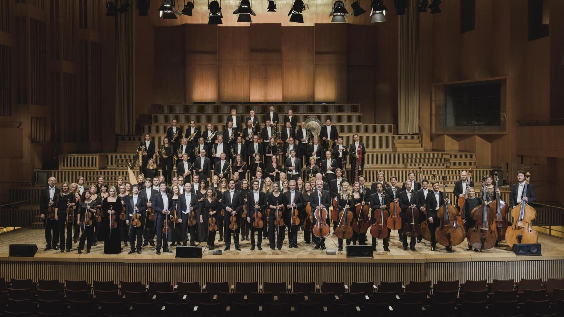 Auf einer Konzertbühne stehen Orchestermitglieder vor einem leeren Publikums-Saal, die ihre Instrumente in der Hand halten.