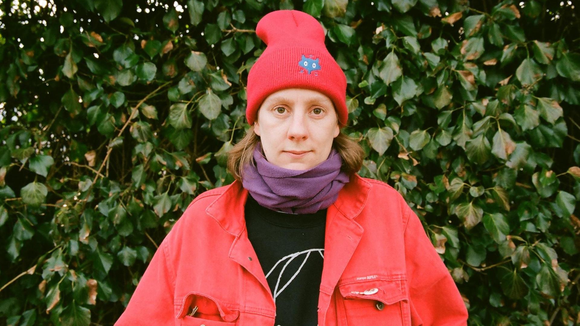 Autorin Anna Haifisch mit einer roten Jacke und roter Wintermütze vor Efeu, guckt direkt in die Kamera