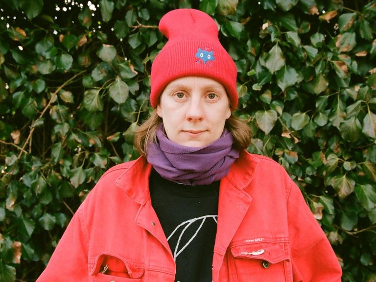 Autorin Anna Haifisch mit einer roten Jacke und roter Wintermütze vor Efeu, guckt direkt in die Kamera