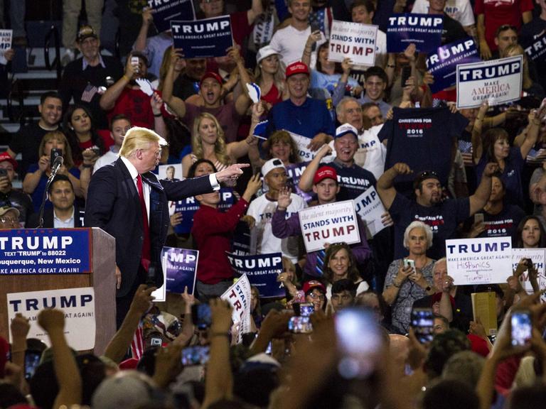 Donald Trump steht bei einer Wahlkampfveranstaltung im Mai 2016 am Rednerpult, umringt von Anhängern, die Schilder mit Trump-Slogans in die Höhe halten.
