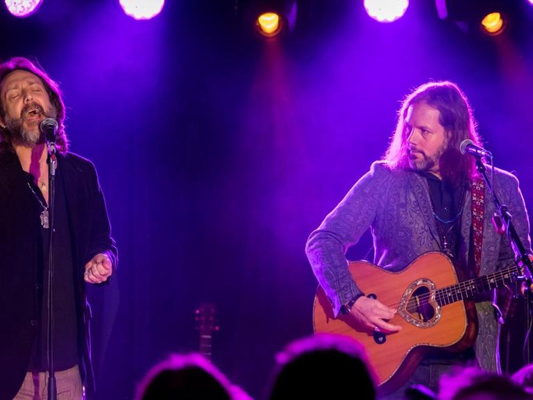 Zwei langhaarige Männer stehen auf einer Bühne. Einer singt in ein Mikrofon, der andere (rechts) spielt Gitarre.