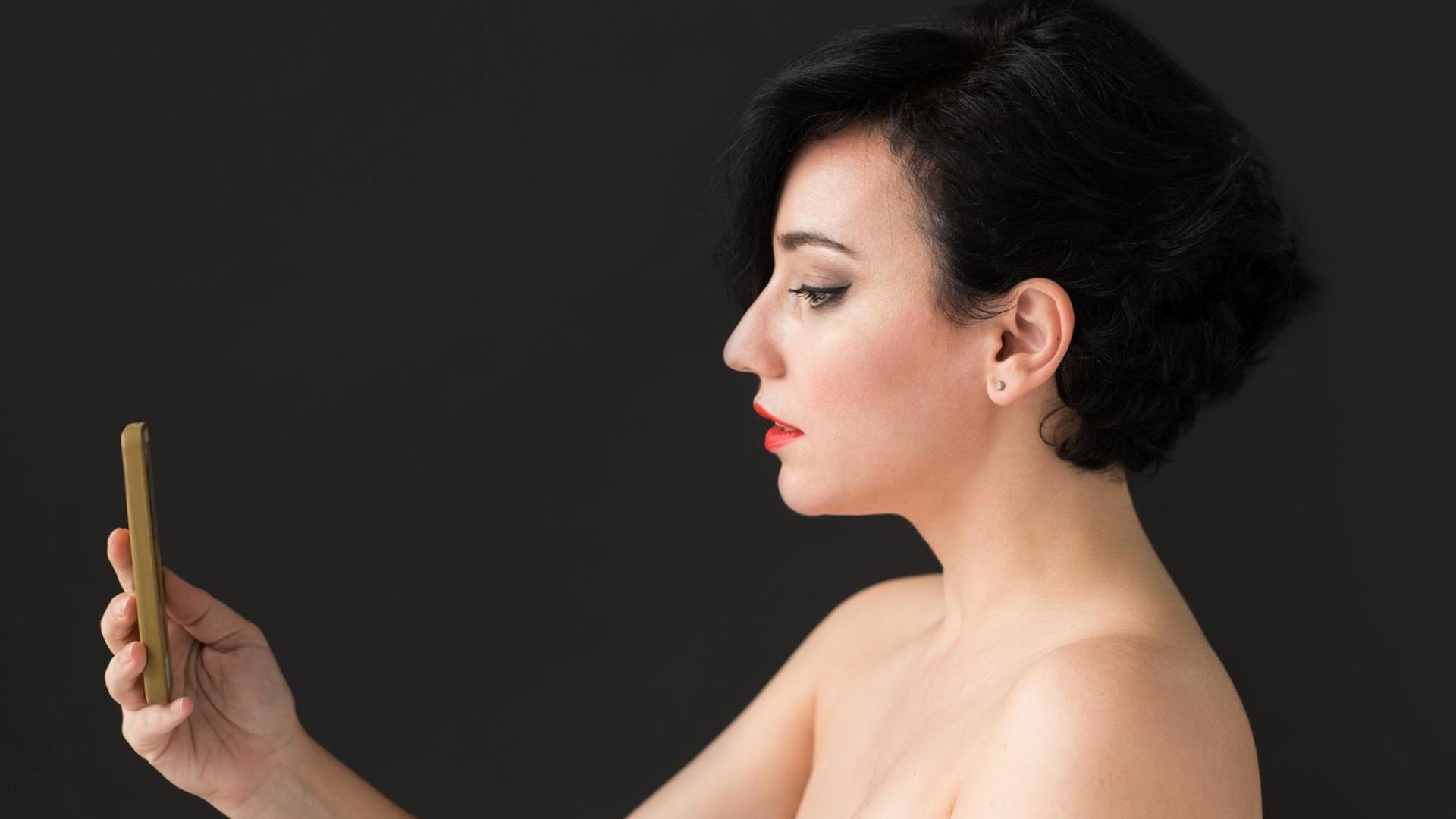 Ein Frau mit nackten Schultern macht ein Selfie von sich. Der Hintergrund ist schwarz.