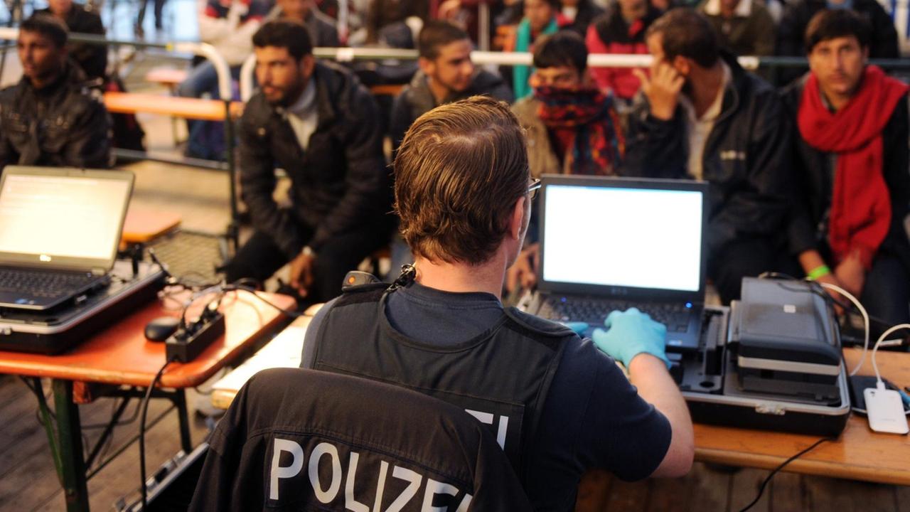 Ein Polizist sitzt in einem Zelt vor einem Computer, Flüchtlinge warten auf ihre Registrierung