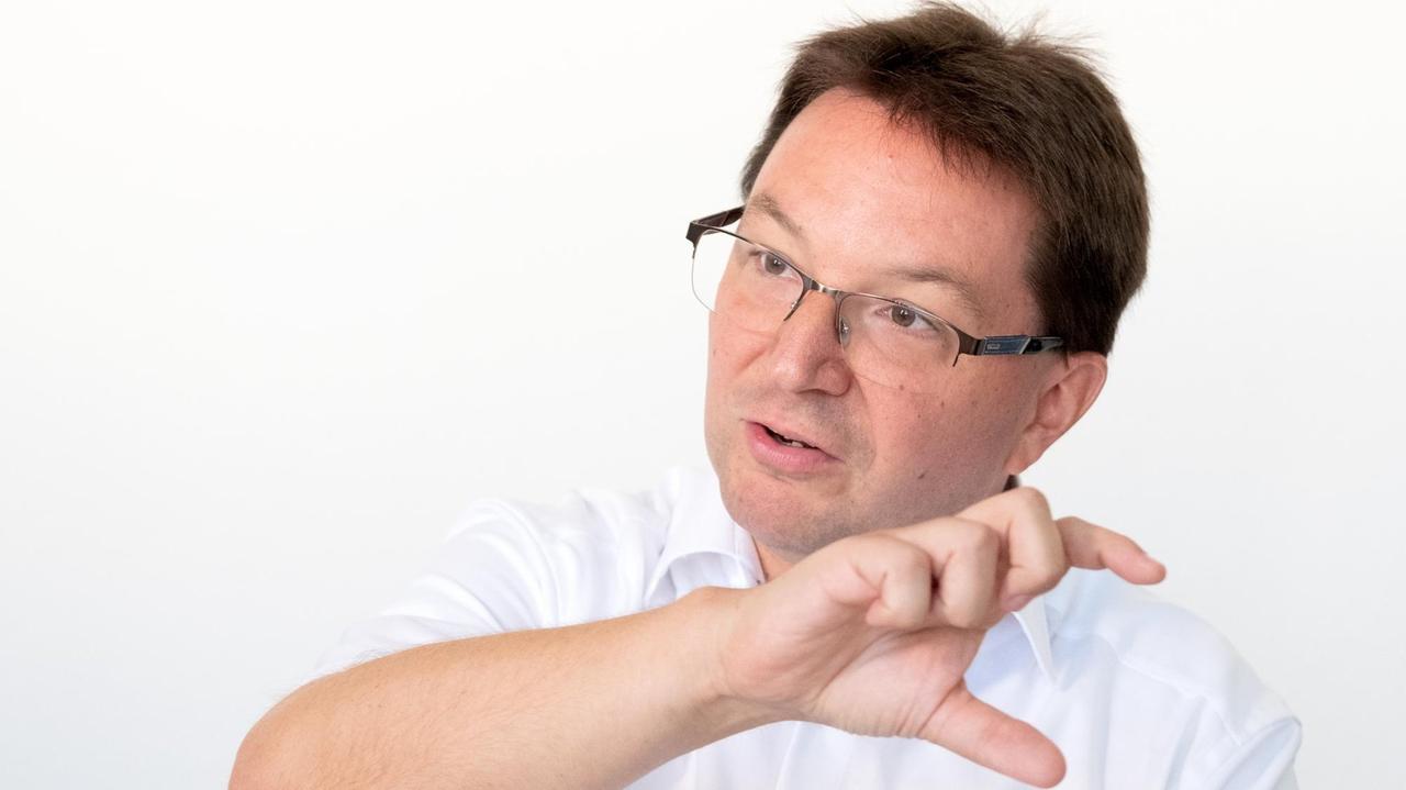 Dr. Michael Blume, der Antisemitismusbeauftragte des Landes Baden-Württemberg, sitzt in einem weißen Hemd vor weißem Hintergrund und gestikuliert mit der rechten Hand.