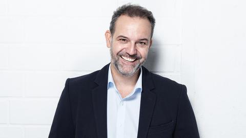 Ein Porträt des Comedians René Steinberg, er steht in einem dunklen Anzug vor einer weißen Wand