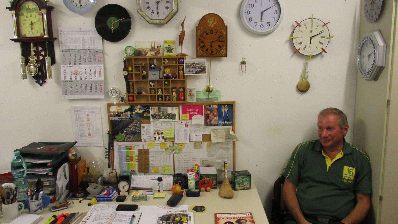 Hausmeister Uwe Mauksch sitzt im Hauswartraum der WG Aufbau in Dresden neben einem Schreibtisch mit allerhand Utensilien und einer ganzen Reihe Uhren an der Wand.