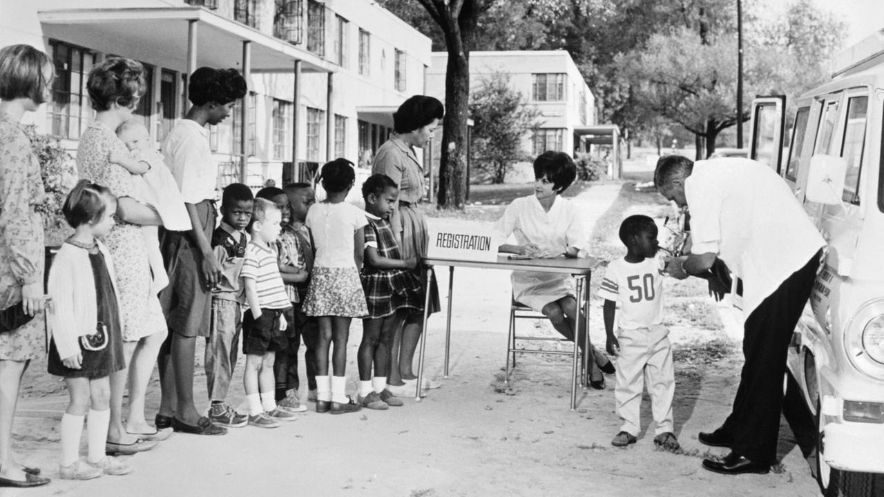 USA 1972: Ein kleiner schwarzer Junge bekommt eine Impf-Injektion. In einer Reihe daneben warten schwarze und weiße Kinder, begleitet von ihren Müttern. Die Szene wurde in der fiktiven "Dixon Tiller County" verortet, einer Modellgemeinde der US-Gesundheitsbehörden - und wurde als Lehrmaterial für Impfaufklärung verwendet