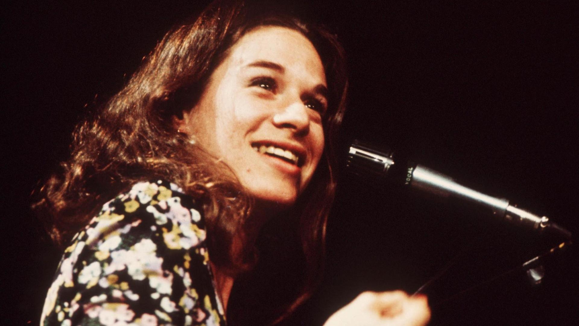 Porträt einer jungen Frau mit langen braunen gewellten Haaren und schwarz-weiß-gelb bedruckter Bluse. Sie strahlt über das ganze Gesicht - vor ihr ein Mikrofon. Vermutlich handelt es sich um eine Bühnensituation, der Hintergrund ist schwarz.