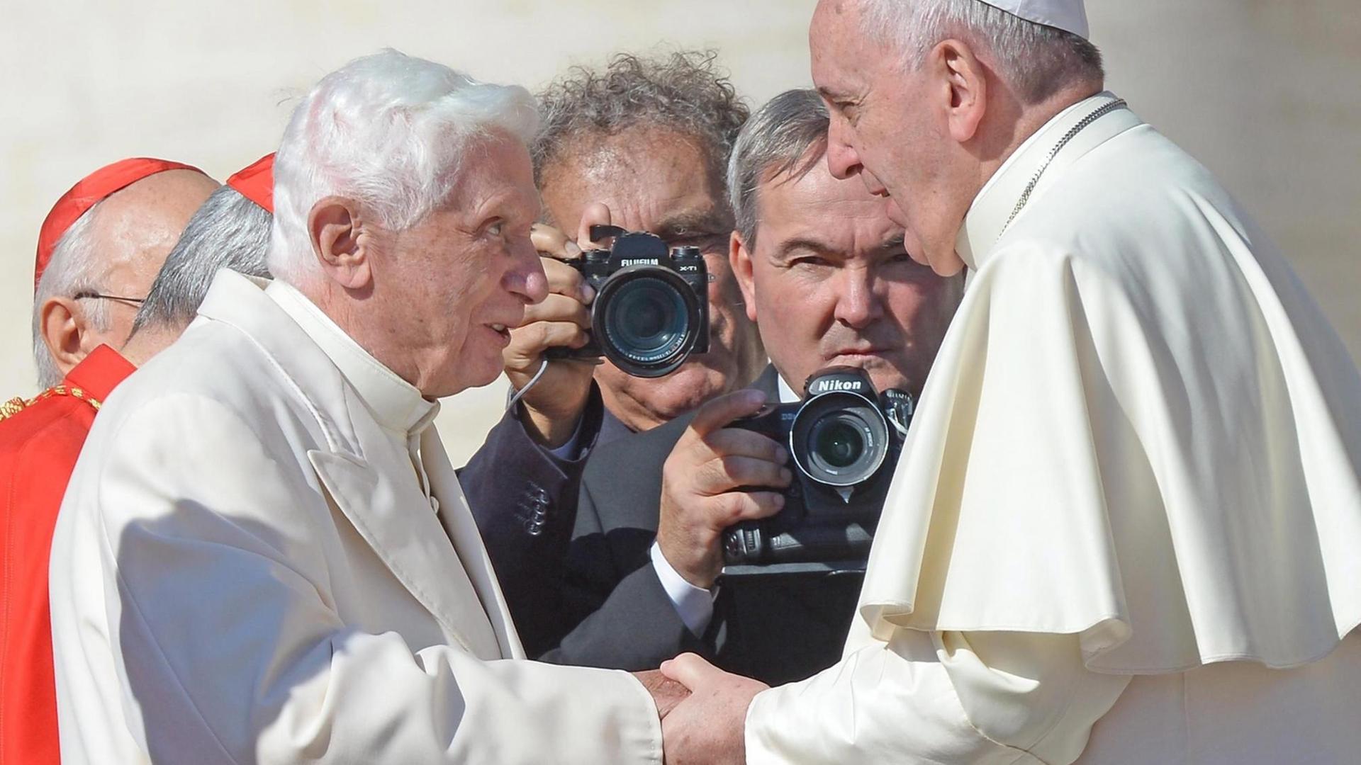 Papst Franziskus und Papst Benedikt XVI. geben einander die Hand. Um sie herum stehen Fotografen.