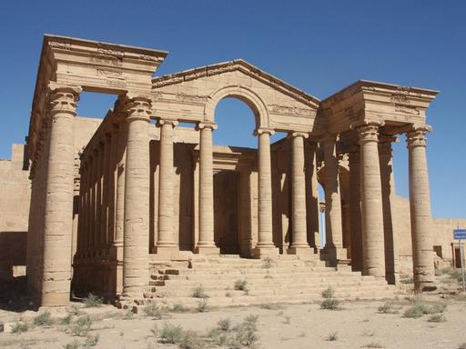 Ein antiker Tempel mit zwei Seitenflügeln und einer Treppe ohne Dach steht in der Wüste.