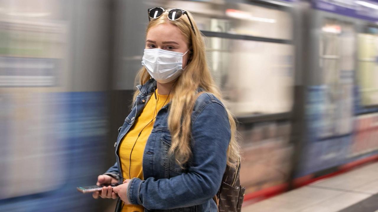 Eine Frau mit einem Mund-Nase-Schutz steht vor einer einfahrenden U-Bahn.