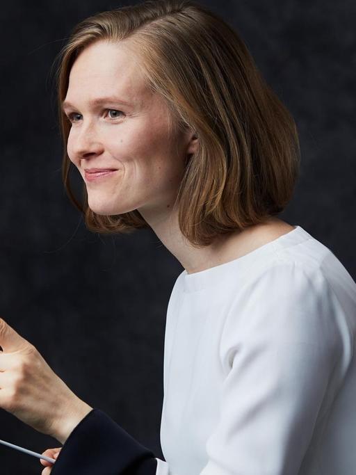 Eine junge Frau blickt freundlich und hebt den linken Zeigefinger. In der anderen Hand hält sie einen Dirigierstab.