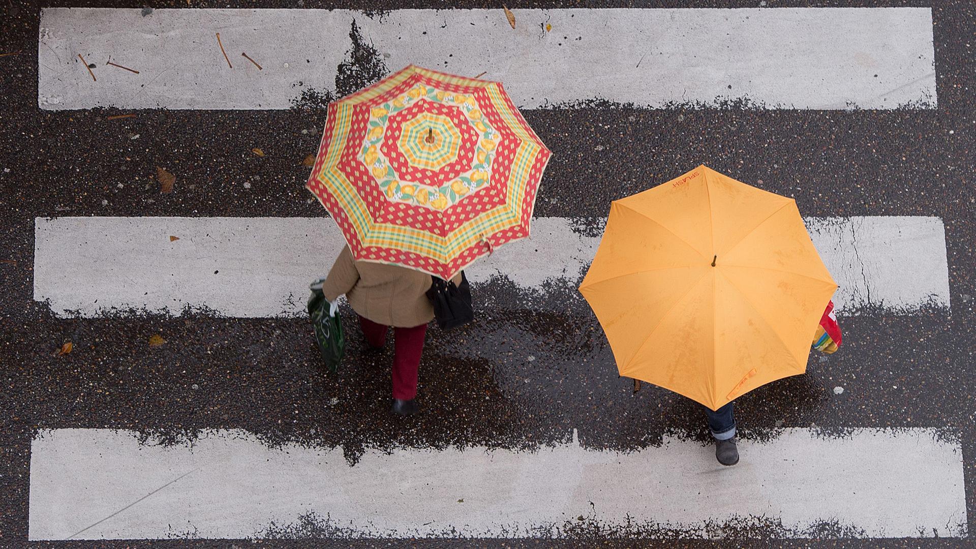 Zwei Passanten mit farbigen Regenschirmen gehen in Stuttgart über einen Zebrastreifen.