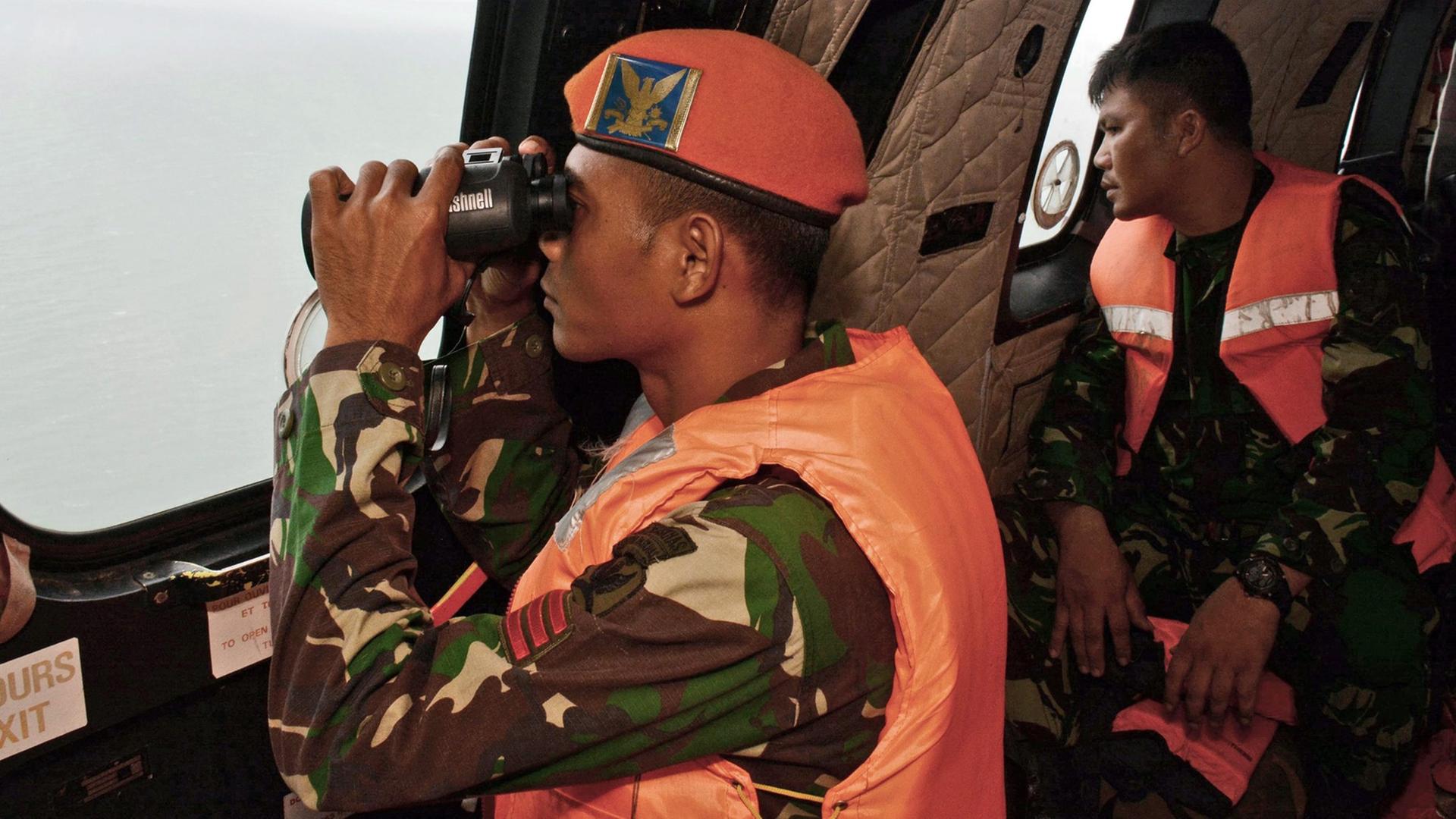 Ein indonesischer Soldat in Uniform und Schwimmweste blickt mit einem Fernrohr aus dem Fenster eines Hubschraubers. Ein indonesischer Soldat in Uniform und Schwimmweste blickt mit einem Fernrohr aus dem Fenster eines Hubschraubers.