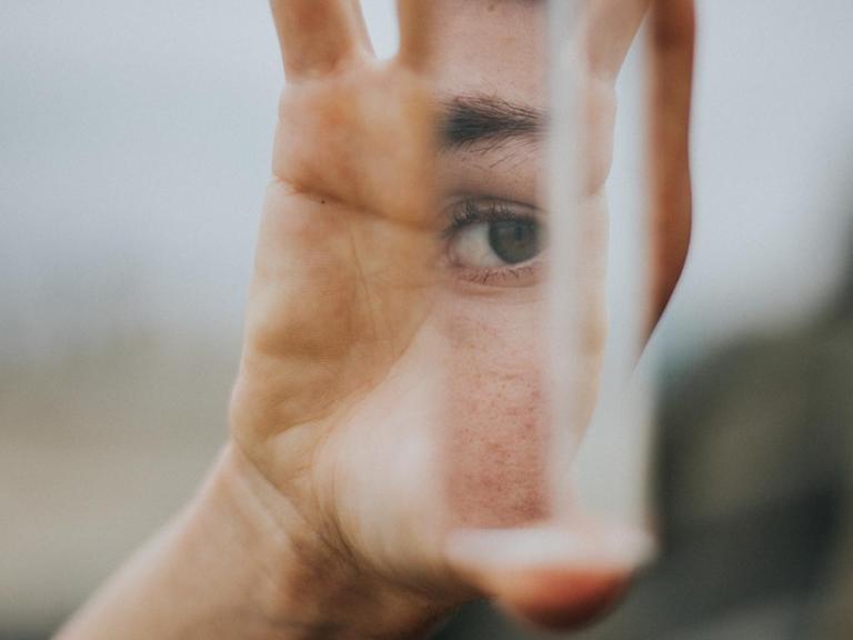 Eine Hand hält einen Spiegel, in dem der Auschnitt eines Gesichts mit einem Auge zu sehen ist.