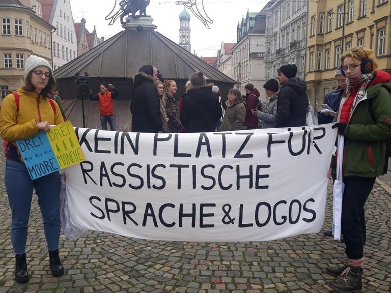 Zwei Menschen halten in Augsburg bei einer Kundgebung zur Umbenennung des Hotels "Drei Mohren" ein Banner mit der Aufschrift "Kein Platz für rassistische Sprache & Logos" zwischen sich, im Hintergrund weitere Personen und ein Brunnen.