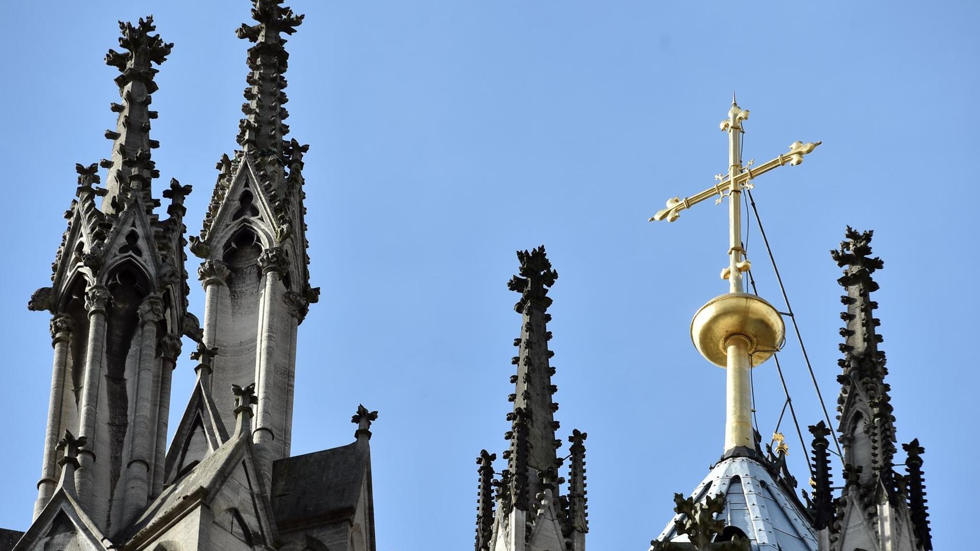 Das Bild zeigt die Spitzen vom Dom in Köln. Ein goldenes Kreuz leuchtet in der Sonne. 