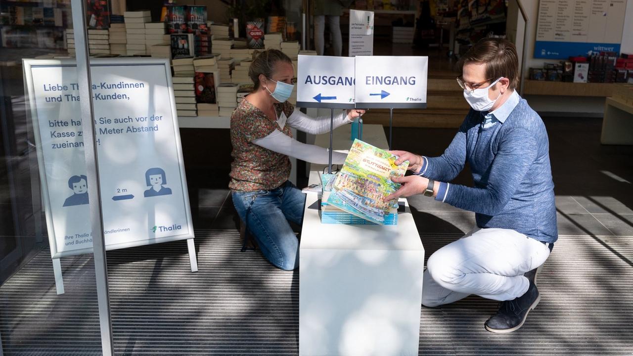 Ein Mann und eine Frau tragen Schutzmasken aus Stoff vor dem Gesicht und platzieren Bücher auf dem Podest vor dem Eingang einer Buchhandlung. Ein Schild hinter ihnen weist Kunden darauf hin, dass sie beim Betreten des Geschäfts voneinander Abstand halten sollen.