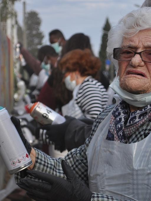 Die 100-Jährige Dona Chica trägt einen Plastikkittel und steht mit einer Sprühdose vor einer Wand. Sie nimmt an einem Graffiti-Kurs für Rentner teil.