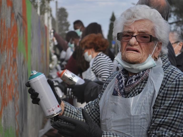 Die 100-Jährige Dona Chica trägt einen Plastikkittel und steht mit einer Sprühdose vor einer Wand. Sie nimmt an einem Graffiti-Kurs für Rentner teil.