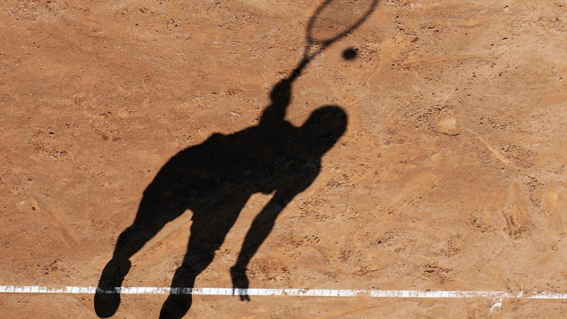 Der Schatten eines Tennisspielers beim Aufschlag auf einem Sandplatz.