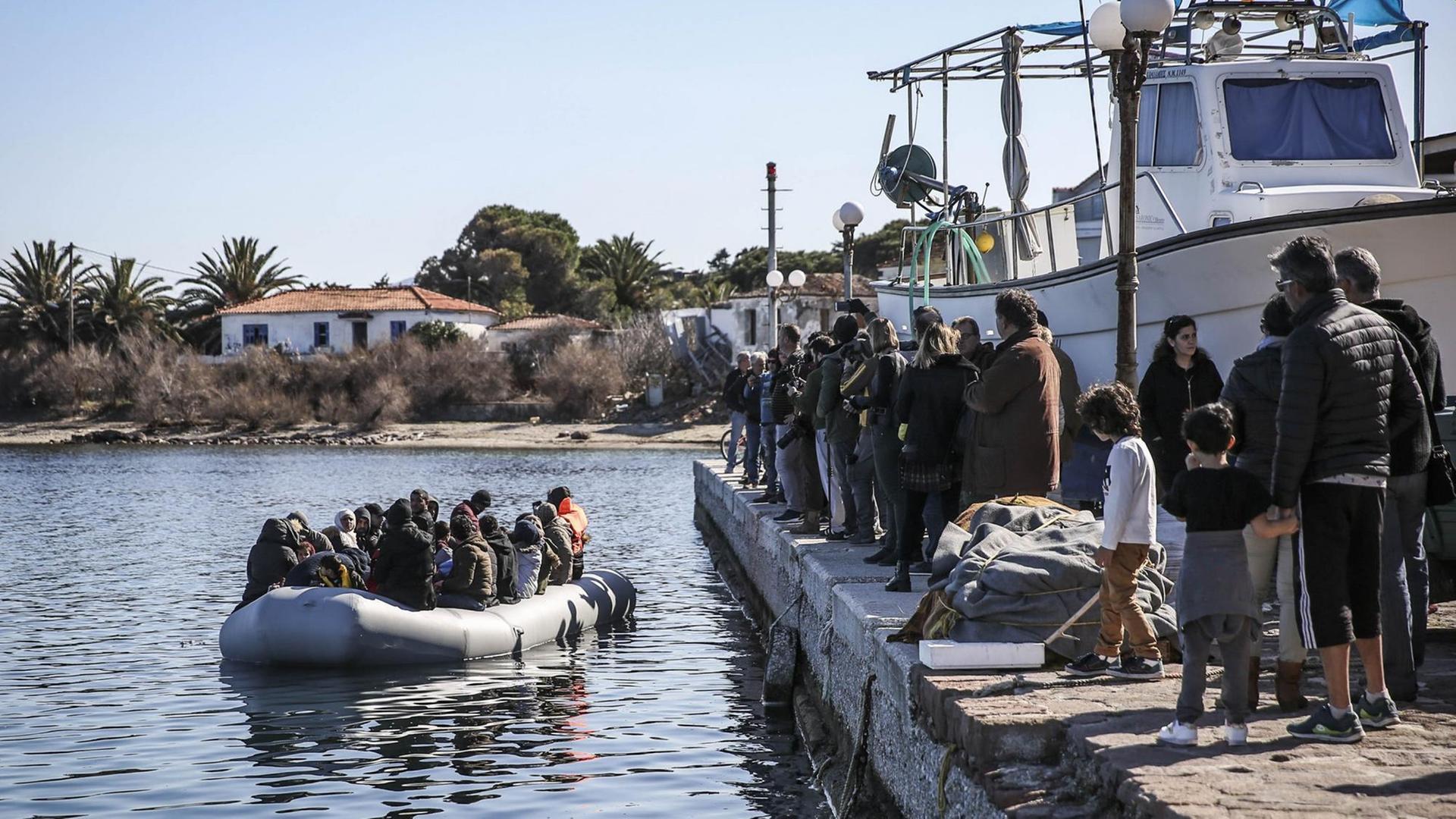 Ein Schlauchboot mit ca. 20 Menschen liegt vor einem Anlegepier, auf dem ähnlich viele Menschen aufgereiht stehen.