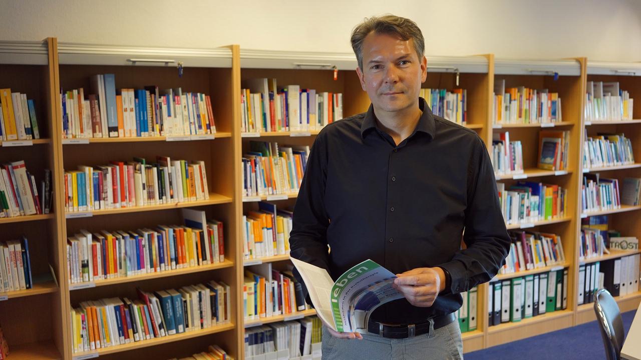 Frank Weise, Geschäftsführer der Krebsgesellschaft Sachsen-Anhalt, steht mit einem Buch in der Hand vor einem Bücherregal.