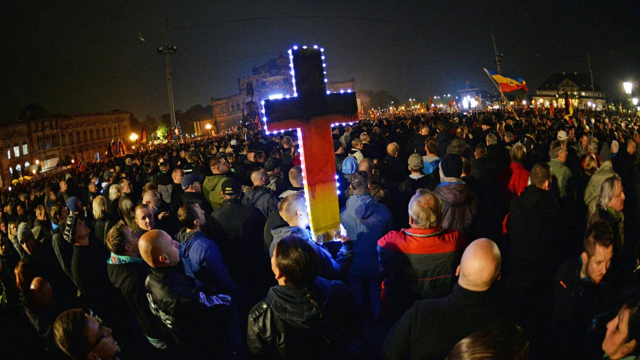 Tausende Anhänger des Bündnisses Pegida stehen auf dem Theaterplatz in Dresden, in der Mitte ist ein in Schwarz-Rot-Gold angemaltes Kreuz zu sehen. aufgenommen am 19.10.2015 