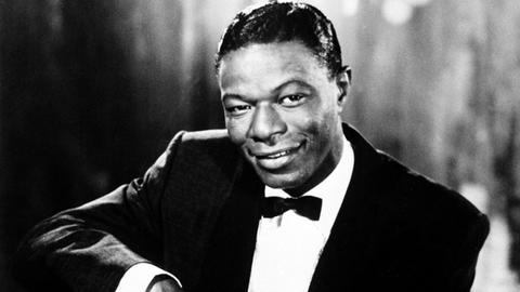 Auf dem s/w Bild ist ein Mann in festlicher Kleidung zu sehen, der auf einem Stuhl sitzt. Er lächelt in die Kamera.