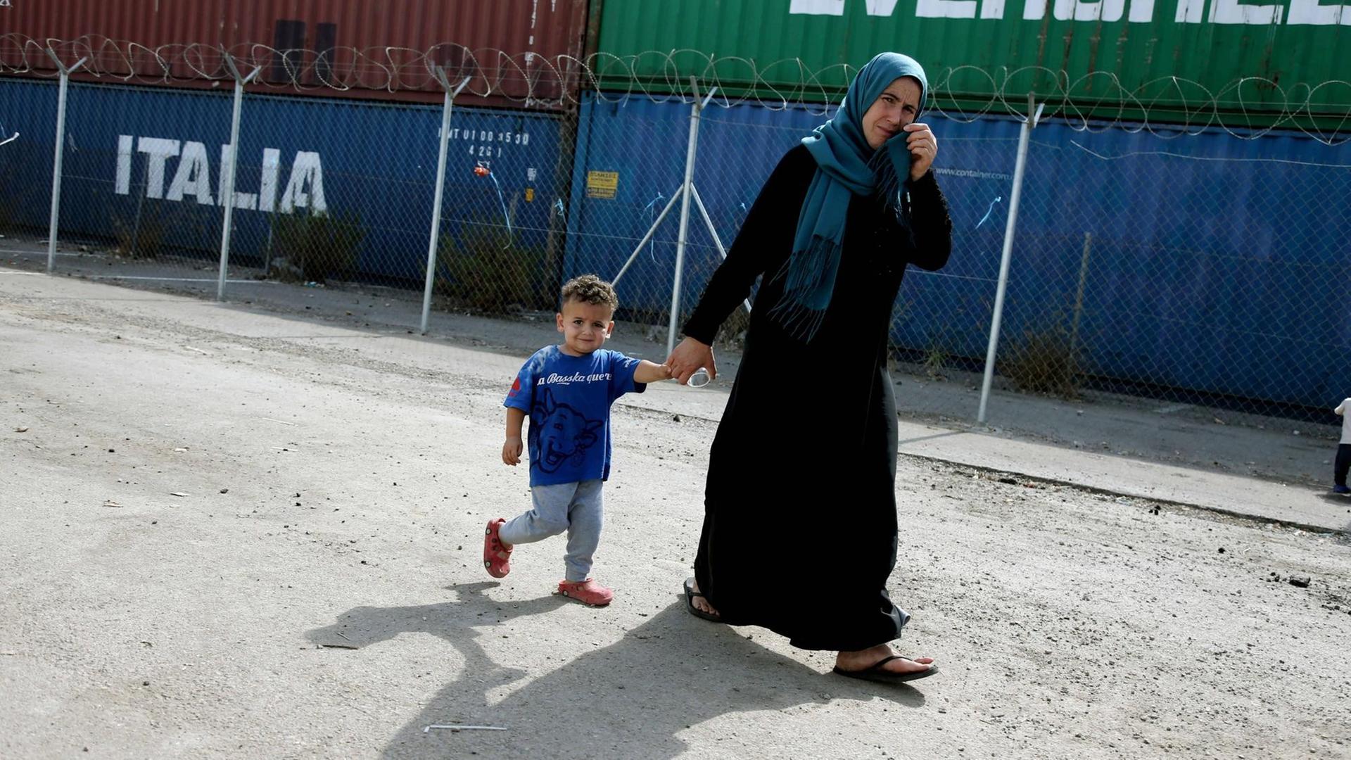 Eine Mutter mit ihrem ca. dreijährigen Sohn läuft vor einem Containerdorf vorbei und schaut in die Kamera. Die Frau trägt ein Kopftuch.