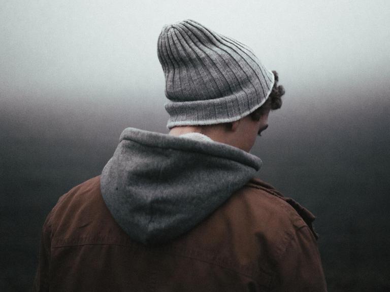 Ein junger Mensch mit Wollmütze, Kapuzenpulli und brauner Jacke schaut vor einem düsteren, nebligen Hintergrund zu Boden.
