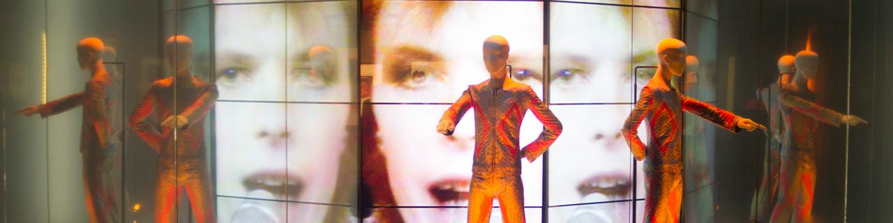 Vor mehreren Bildschirmen, die David Bowie zeigen, stehen Schaufensterpuppen, die glitzernde Anzüge tragen.