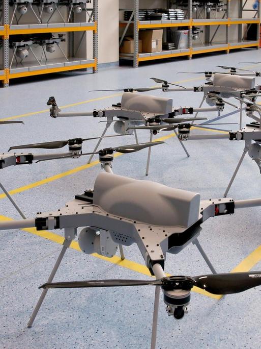 Aufgereihte Drohnen stehen nebeneinander auf einem blauen Linoliumboden in einer Lagerhalle.