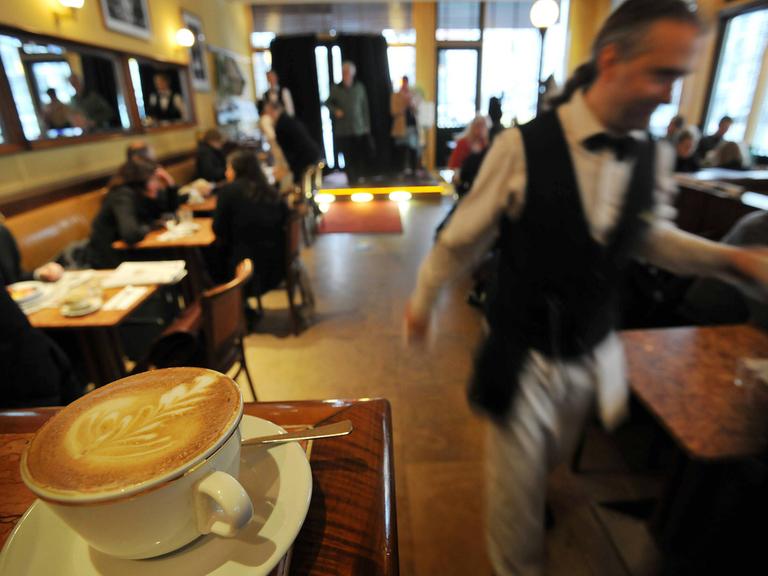 Blick in ein Restaurant, auf einer Theke im Vordergrund steht ein Kaffee, ein Kellner läuft durchs Bild.